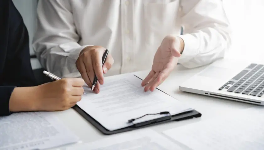 Deux personnes discutent d'un document, l'une signant, l'autre expliquant. Une commission d'intervention est en cours.