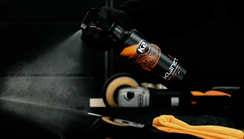 Rękawiczka w czarnej rękawiczce rozpyla produkt K2 KLINET na czarny lakier samochodowy. W tle polerka.