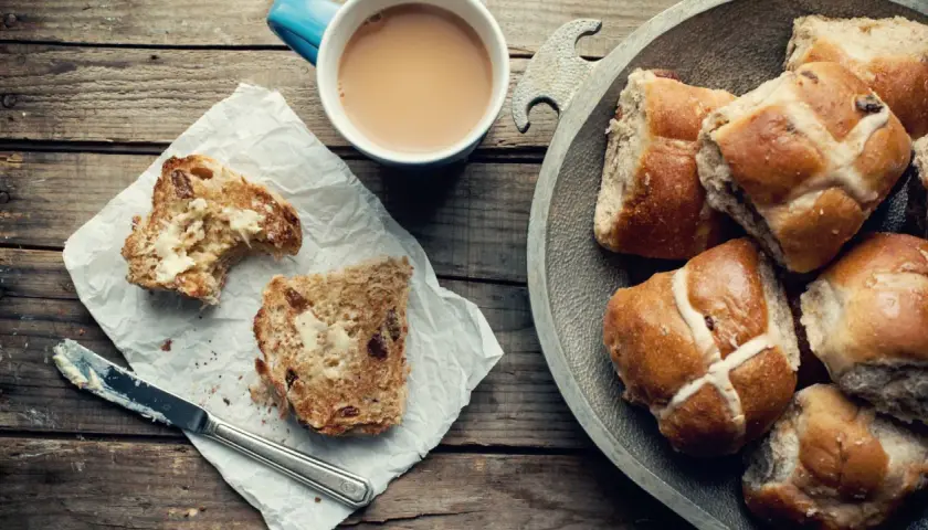 Na drewnianym stole leżą świeże, pachnące hot cross buns, obok kubek kawy i kromka posmarowana masłem.
