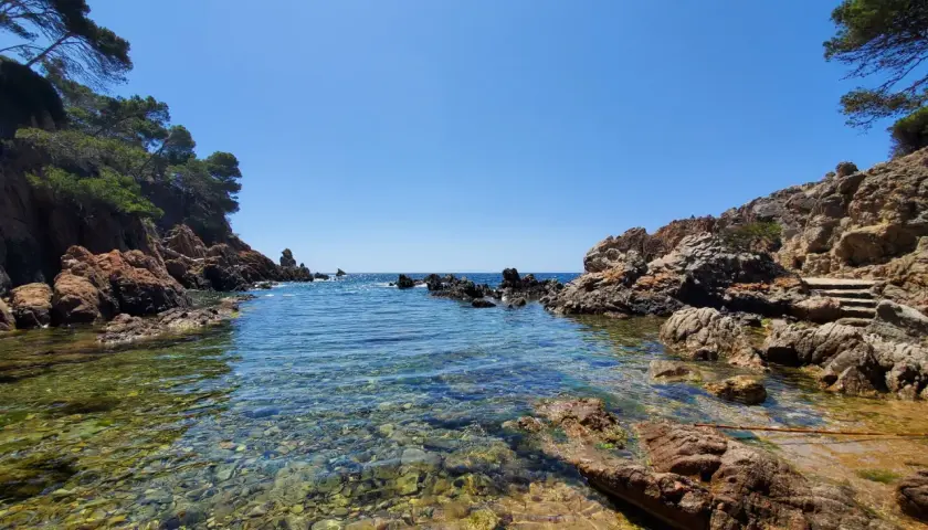 Aguas cristalinas y rocas rojizas en una cala idílica, un tesoro entre las mejores calas de la Costa Brava.
