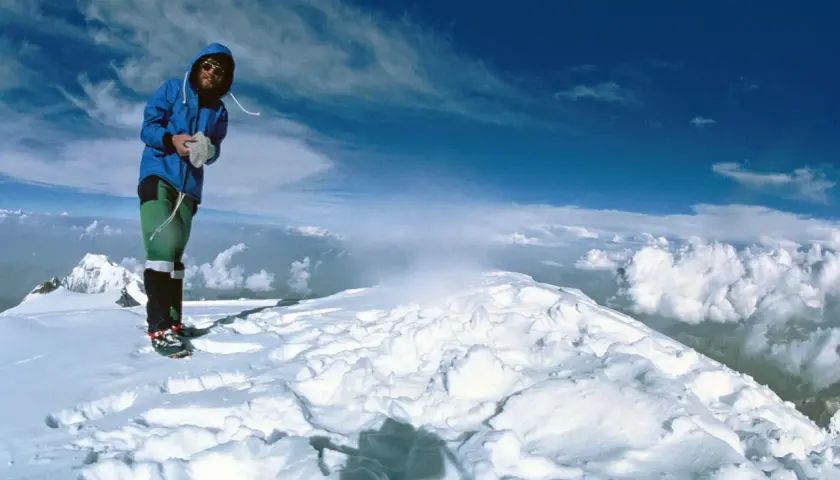 Reinhard Messner steht auf einem schneebedeckten Gipfel, umgeben von Wolken und blauem Himmel.