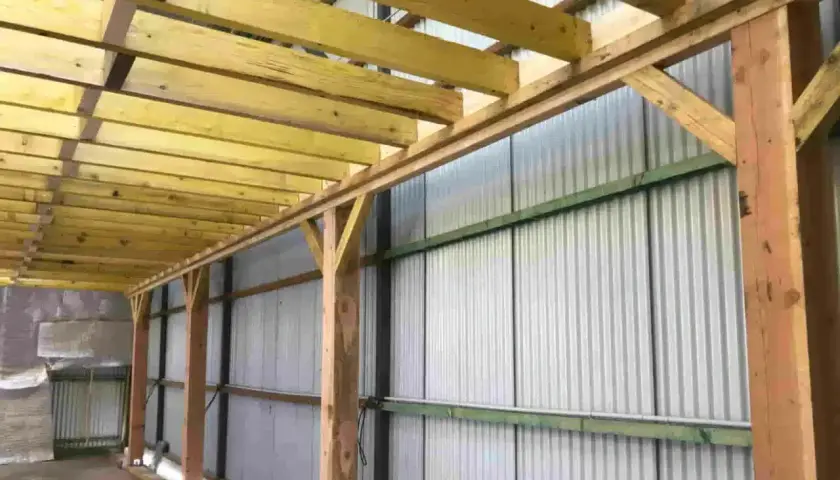 Structure en bois avec madriers ou bastaings soutenant un toit, le long d'un mur en tôle ondulée.