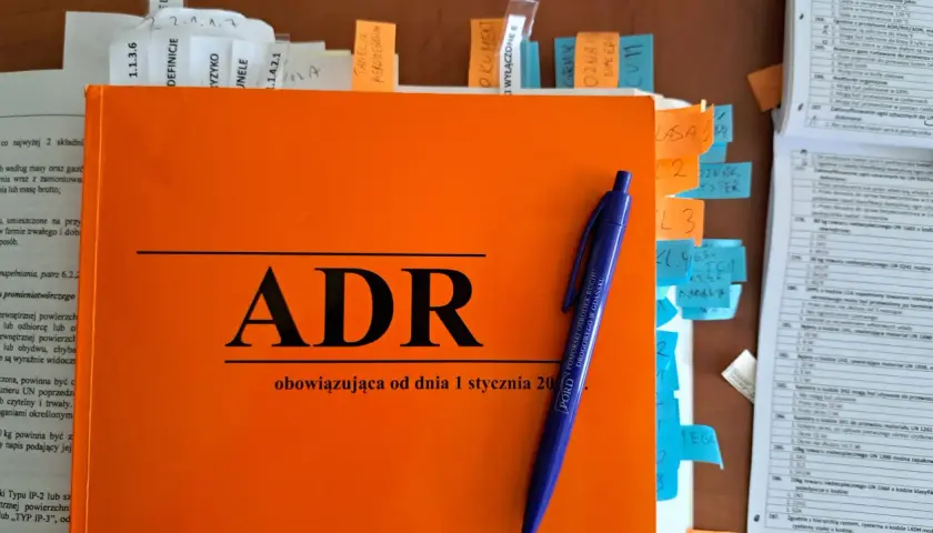 Pomarańczowa książka z napisem "ADR" i niebieski długopis. To materiały do kursu **doradca ADR**.