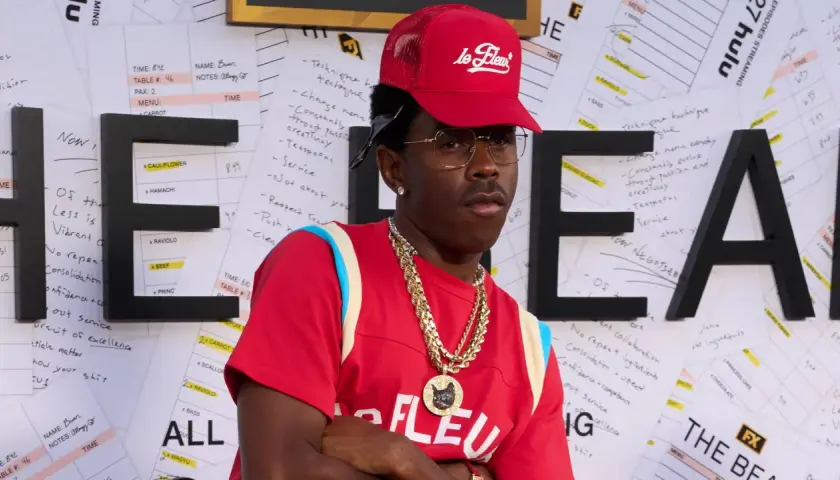 Tyler, The Creator w czerwonej czapce i naszyjnikach, na tle liter "HE" i notatek.