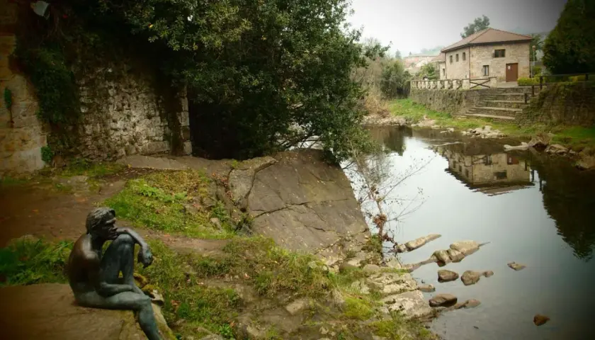 Escultura de hombre pensativo junto a un río, con una casa de piedra reflejada en el agua. Un lugar tranquilo que ver cerca de Liérganes.
