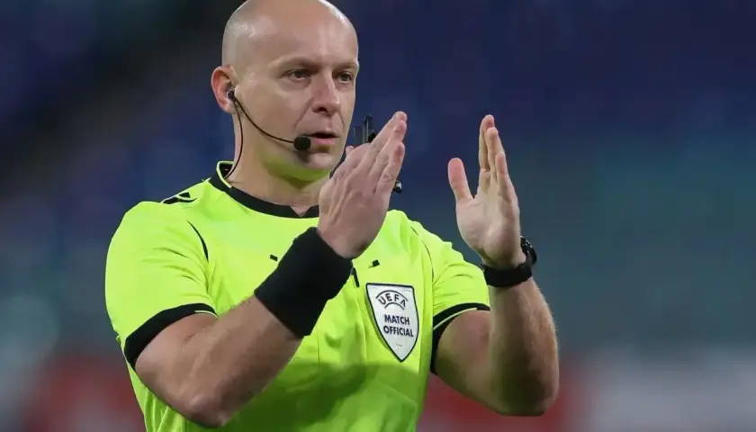 Sędzia piłkarski w żółtej koszulce z emblematem UEFA, z mikrofonem przy uchu, pokazuje gest rękami. Ciekawe, ile zarabia sędzia na takim meczu.