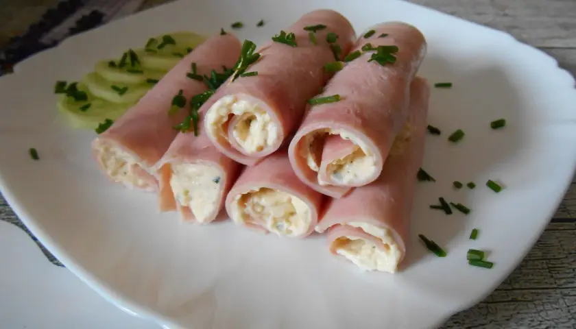Roladki z twarogiem, zawinięte w plastry szynki, ozdobione szczypiorkiem i podane z plasterkami ogórka.