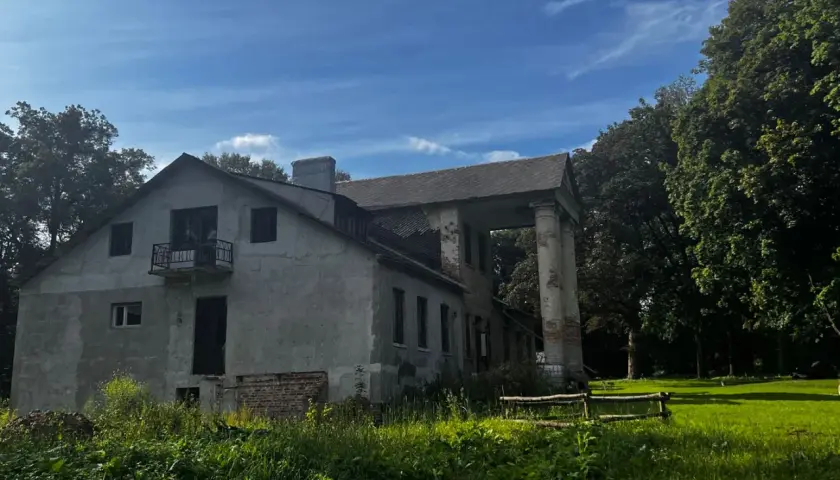 Zniszczony dworek z kolumnami i balkonem, otoczony zielenią.