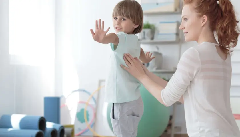 Ein Kind mit Skoliose erhält osteopathische Behandlung. Eine Therapeutin legt sanft die Hände auf den Rücken des Jungen, der sich leicht wegdreht.