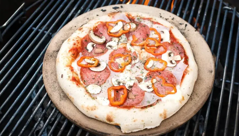 Eine köstliche Pizza mit Salami, Pilzen und Paprika wird auf einem Pizzastein über glühenden Kohlen auf dem Grill zubereitet.
