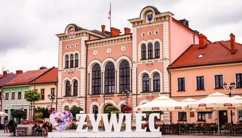 Duże białe litery "ŻYWIEC" na rynku, obok serca z ptaszkiem. W tle różowy ratusz i kawiarniane parasole.