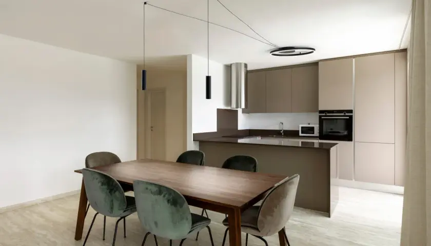 Un lampadario decentrato crea un'atmosfera moderna sopra un tavolo da pranzo in legno con sedie in velluto verde. Soluzioni di illuminazione eleganti per ogni spazio.