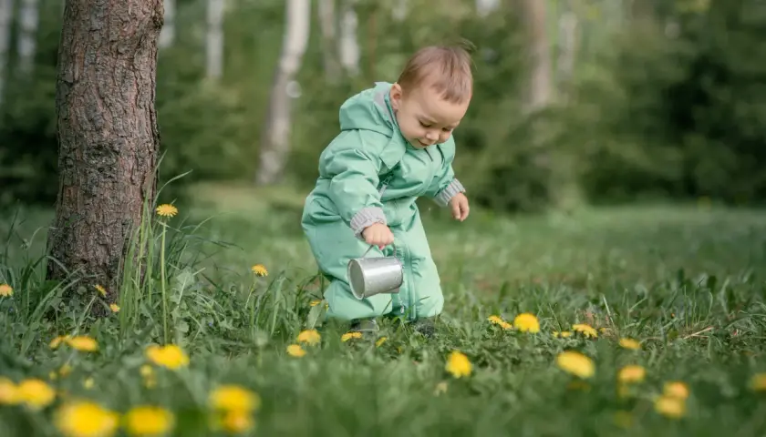 Dziecko w zielonym kombinezonie z konewką w trawie z żółtymi kwiatami.