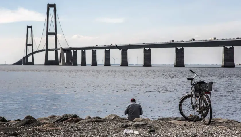 En man sitter vid vattnet med sin cykel, blickandes mot den imponerande Stora Bältbron.