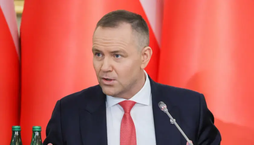 Mężczyzna w garniturze i czerwonym krawacie przemawia na tle czerwonych flag. Dyskutuje, dlaczego prezydent zapowiada veto wobec ustawy o wyborze sędziów do KRS.
