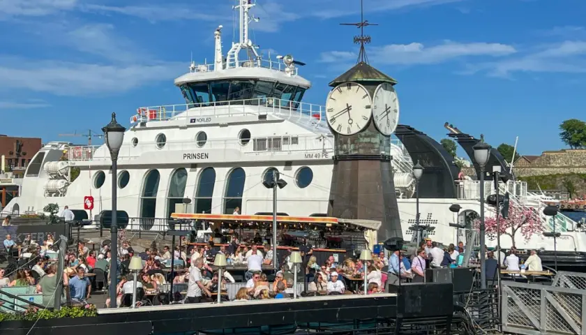 Tętniący życiem deptak w Oslo, z ludźmi siedzącymi w kawiarniach, na tle białego statku i zabytkowej wieży zegarowej. Oslo czy warto? Zdecydowanie tak!