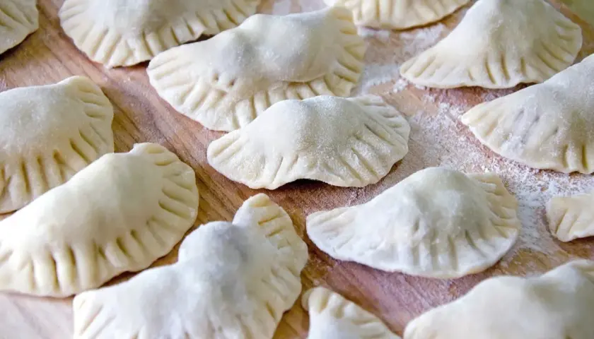 Surowe pierogi z kaszą gryczaną ułożone na drewnianej desce, posypane mąką.