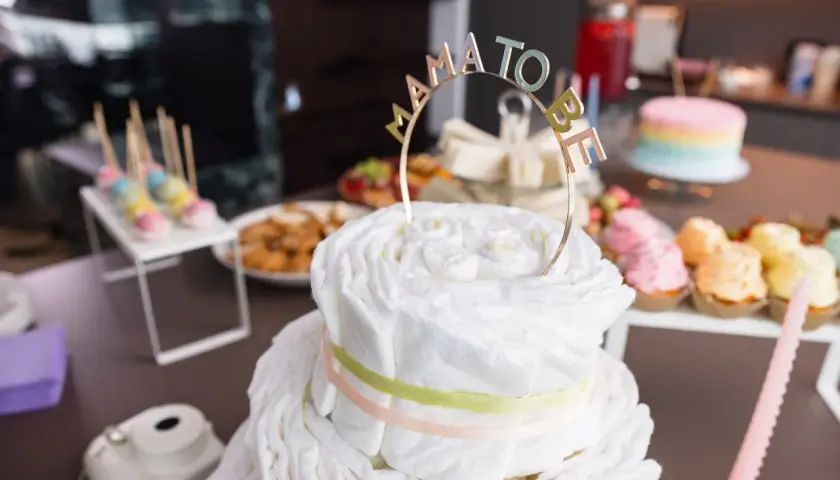 Una torta di pannolini decorata con un topper "Mama to Be" per un baby shower. Sul tavolo anche cupcakes colorati e dolcetti.