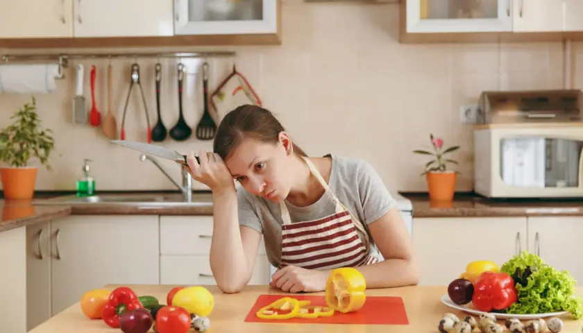 Zmęczona kobieta z nożem w dłoni zastanawia się, czy dieta pudełkowa się opłaca. Na stole warzywa i jajka.