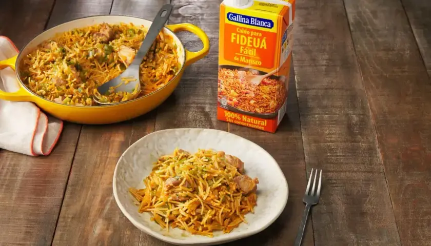 Un plato de fideos a la cazuela servido en una cazuela amarilla y en un plato blanco, junto a un cartón de caldo para fideuá.