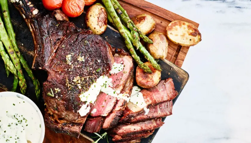 En saftig tomahawk-biff, perfekt grillad, serveras med rostade tomater, sparris och potatis. En krämig sås fulländar denna grillupplevelse.