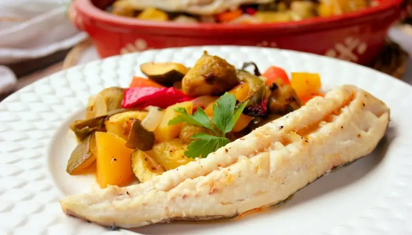 Filete de merluza al horno con verduras asadas, como pimientos, calabacín y patatas. Un plato saludable y delicioso.
