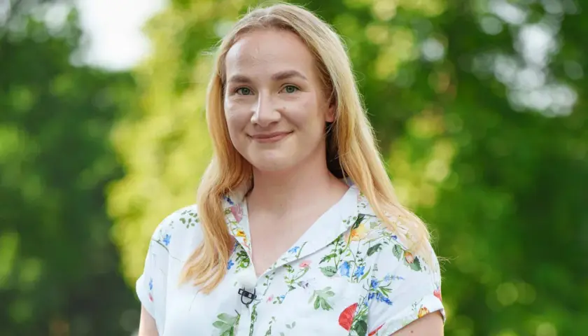 Uśmiechnięta Klaudia, uczestniczka "Rolnik szuka żony", w kwiecistej koszuli na tle zieleni.