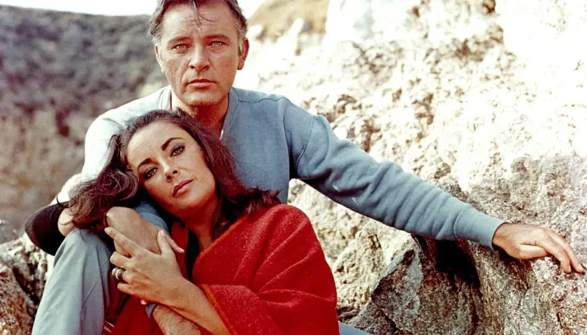 Elizabeth Taylor, żona Richarda Burtona, opiera głowę na jego ramieniu, otulona czerwoną narzutą, siedząc na kamieniach.