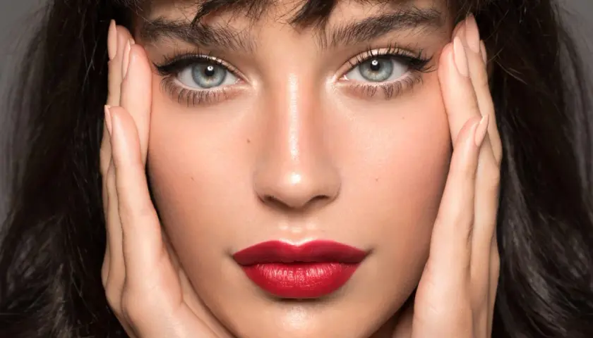 Close-up of a woman's face with perfect winged eyeliner and red lipstick, hands framing her face.