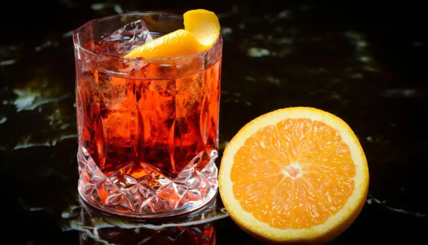 Ein erfrischender Negroni mit Eis und Orangenzeste, daneben eine saftige Orangenscheibe – die perfekten Negroni Zutaten.