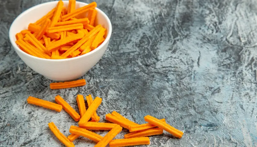 Pomarańczowe paluszki snacków w białej misce i rozsypane obok na szarym tle.