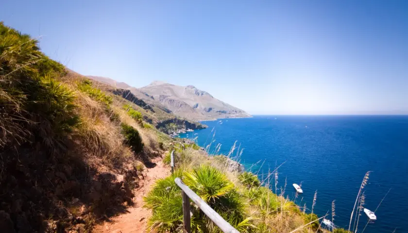 A scenic hiking trail winds through the rugged landscape of Lo Zingaro Nature Reserve, overlooking the brilliant blue Mediterranean Sea dotted with boats.