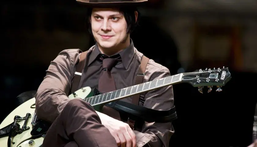 Jack White w brązowym kapeluszu i garniturze gra na gitarze.