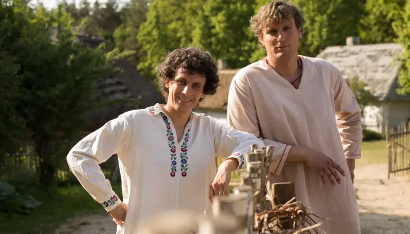 Dwóch mężczyzn w strojach ludowych, jak z filmu "Sami swoi", pozuje na tle wiejskiego krajobrazu.