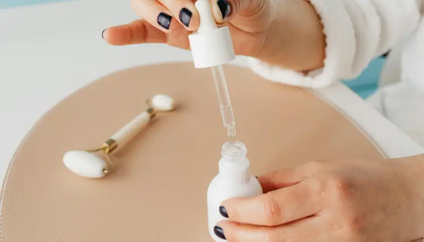 Dłoń z ciemnym lakierem do paznokci trzyma pipetkę z serum, obok jadeitowy roller. Pamiętaj, z czym nie łączyć niacynamidu.