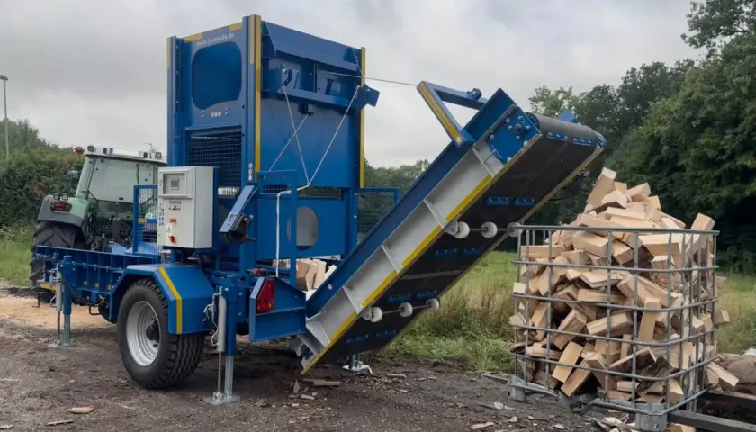 Ein Förderband für Holz transportiert gespaltene Holzscheite in einen Gitterbox-Container. Ein Traktor steht im Hintergrund.