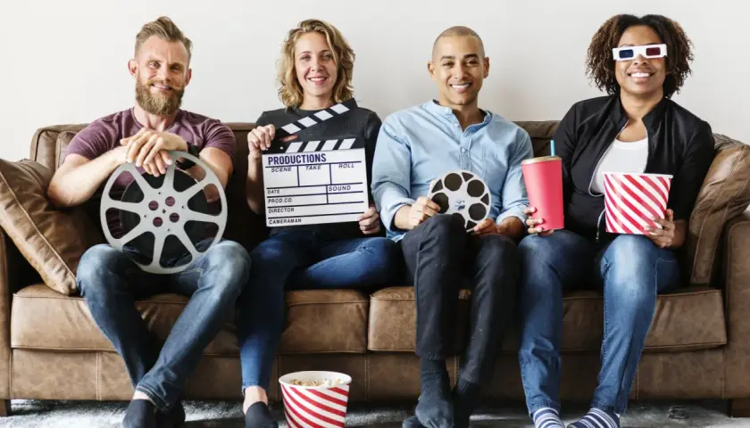 Cztery osoby siedzą na kanapie, trzymając rekwizyty filmowe: klaps, rolki taśmy filmowej, popcorn i napój. Różne rodzaje filmów i ich tworzenie.
