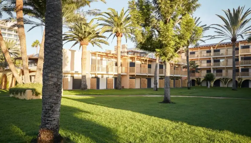 Edificio del Robinson Club Jandia Playa rodeado de palmeras y césped verde, con balcones de madera y fachadas claras.