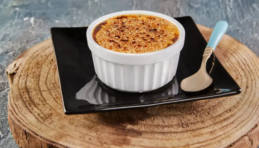 Crème brûlée avec une croûte caramélisée dorée, servie dans un ramequin blanc sur une assiette noire, avec une cuillère à la touche champignon.