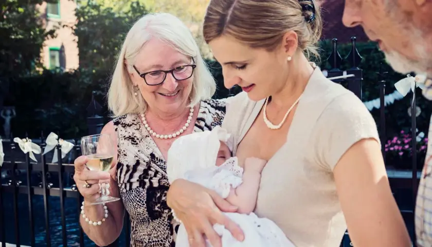 Elegancka mama karmi dziecko na chrzcinach. Babcia z kieliszkiem wina i dziadek patrzą z uśmiechem.