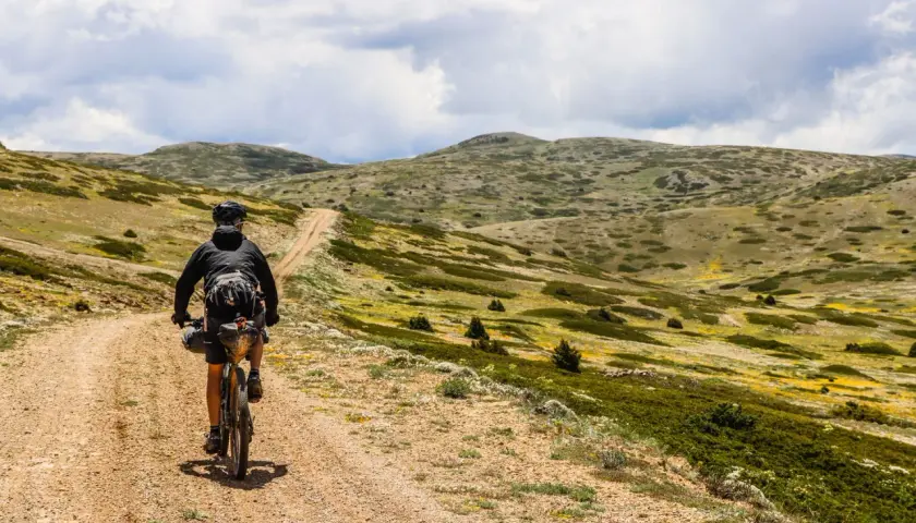 Ciclista pedaleando por un camino de tierra en **montañas vacías** bajo un cielo nublado.
