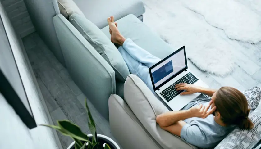 Mężczyzna relaksuje się na kanapie, pracując na laptopie. W tle widać roślinę doniczkową. To idealne miejsce do pracy zdalnej, jak na platformie ozzion.