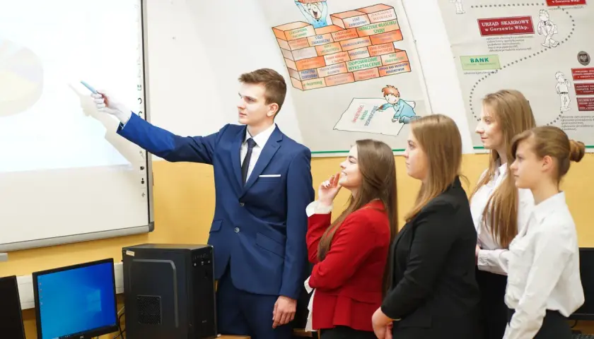 Młody technik ekonomista tłumaczy wykres na tablicy interaktywnej grupie uczniów.