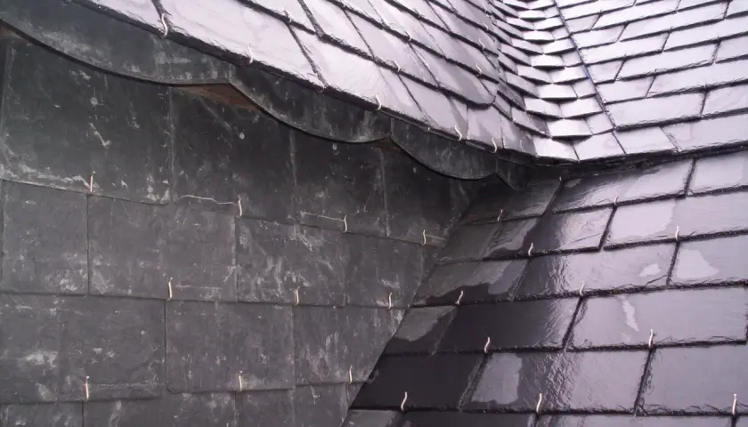 Détail d'une toiture ardoise, avec des ardoises sombres et brillantes, fixées par des crochets métalliques.