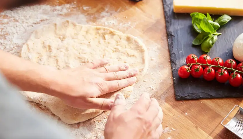 Hände formen den **pizzateig auftauen** auf einer bemehlten Holzoberfläche. Daneben liegen frische Tomaten, Mozzarella und Basilikum.