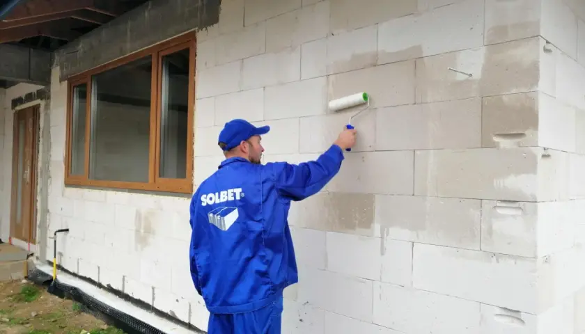 Pracownik w niebieskim uniformie nakłada wałkiem grunt pod styropian na ścianę z bloczków betonu komórkowego.