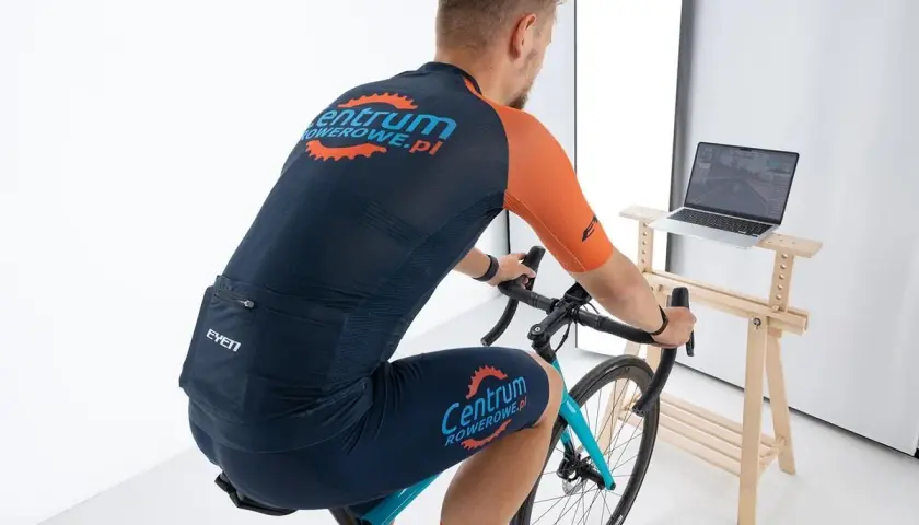 Mężczyzna na rowerze stacjonarnym, ćwiczący cardio. Na ekranie laptopa widać wirtualną trasę kolarską.