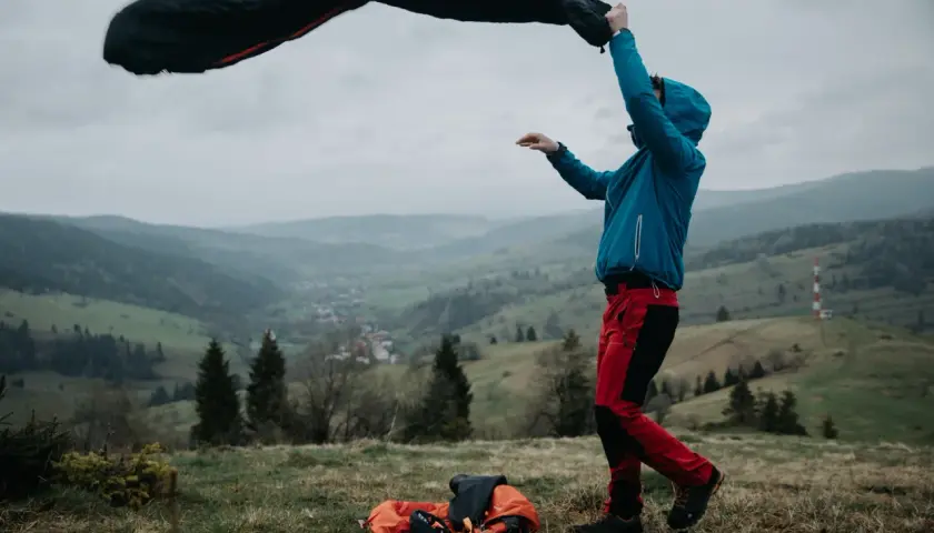 Osoba w niebieskiej kurtce i czerwonych spodniach rozkłada śpiwór puchowy czy syntetyczny na tle górskiego krajobrazu.