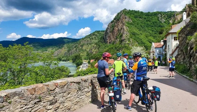 A group of cyclists on a Danube bike tour pauses by a stone wall, with a river and lush green hills in the background.