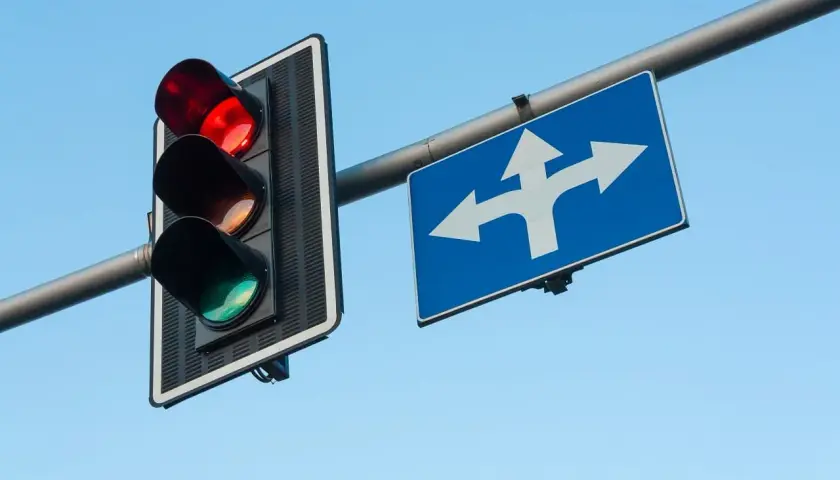 Migające światła na przejeździe kolejowym wskazują, że zbliża się pociąg. Znak pokazuje, że można jechać prosto lub skręcić.